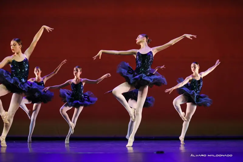 Fünf Ballerinas in marineblauen Paillettentutus balancieren auf einem Bein vor tiefroter Kulisse