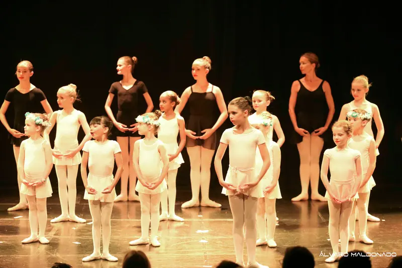 Jeunes élèves de ballet en blanc devant des élèves plus grandes et une professeure en noir, sur une scène à lumière chaude