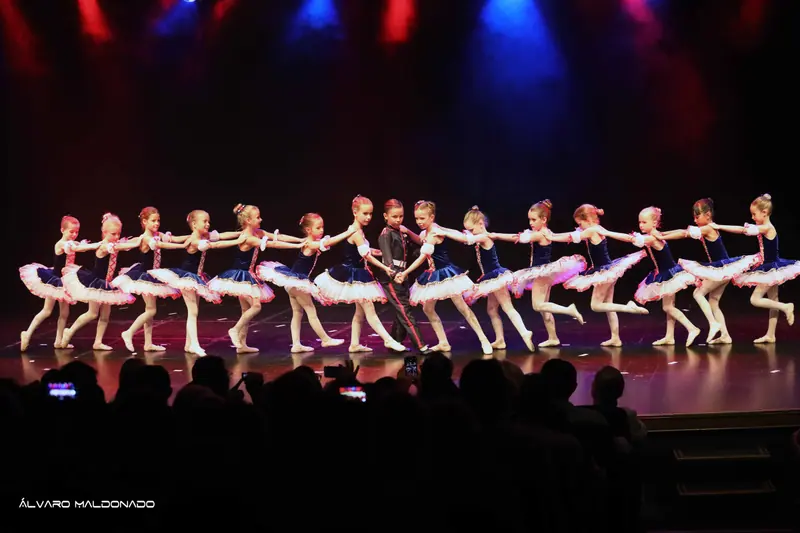 Une longue chaîne de filles en tutus bleu marine et blanc se tient autour d’un garçon en uniforme sous des lumières rouges et bleues