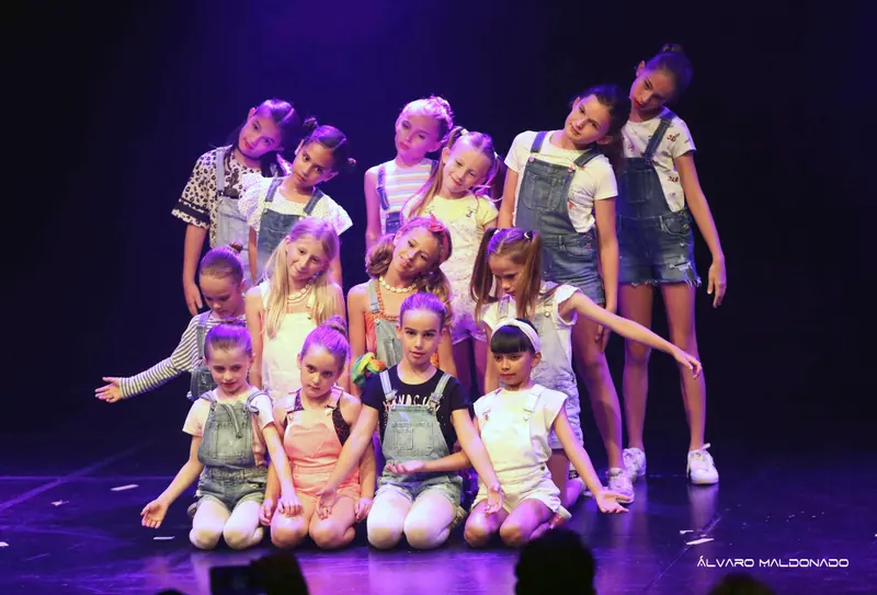 Des filles en salopettes en jean et t-shirts posent en groupe serré, certaines à genoux devant, sous une lumière violette