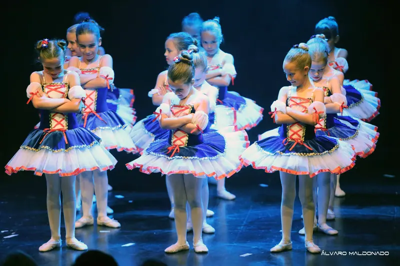 Des filles en tutus bleu marine et blanc à laçage rouge, bras croisés et tête baissée, sourient timidement sur scène