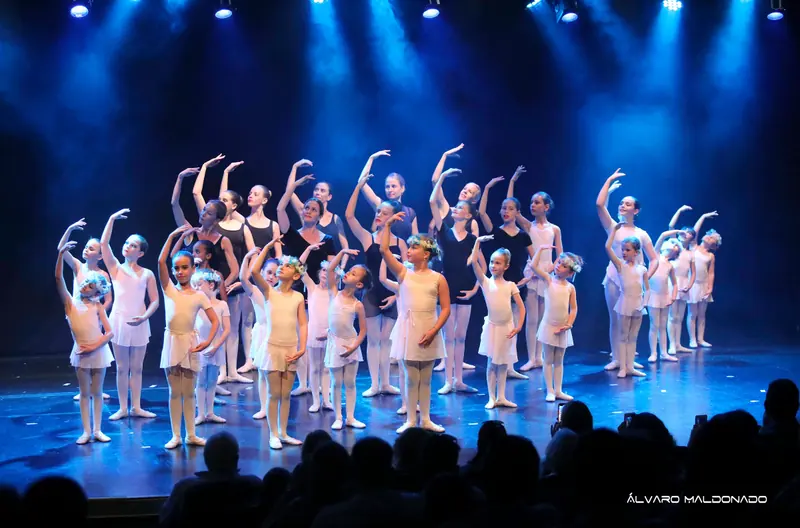 Un grand groupe de ballet en blanc et noir, un bras arrondi au-dessus de la tête, sous des projecteurs bleus face au public