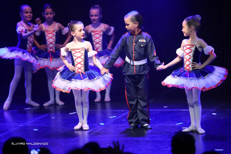 Un garçon en uniforme de soldat tient la main de deux filles en tutus bleu marine et blanc, d’autres regardent derrière