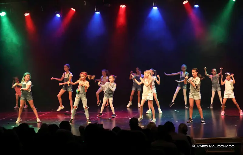 Des filles en salopettes en jean et baskets dansent une chorégraphie street jazz sous des projecteurs rouges, bleus et verts