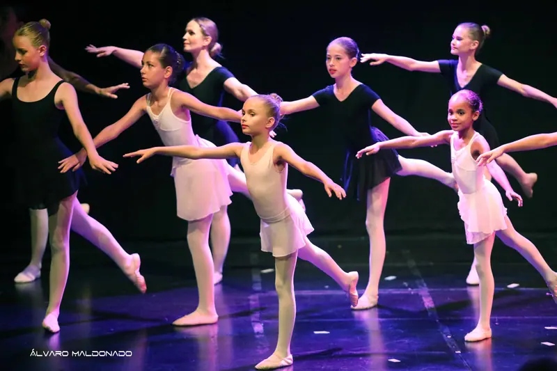 Élèves de ballet en justaucorps blancs et noirs en arabesque, bras tendus, sous une lumière rose