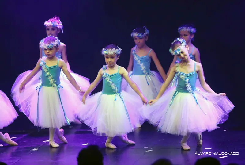 Des filles en corsage turquoise, longs tutus blancs et couronnes de fleurs tiennent leur jupe et tendent un pied, lumière violette