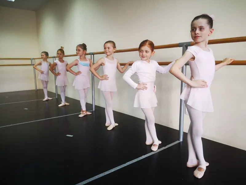 Jeunes élèves de ballet en justaucorps et jupettes rose pâle à la barre, mains sur les hanches, dans un studio lumineux