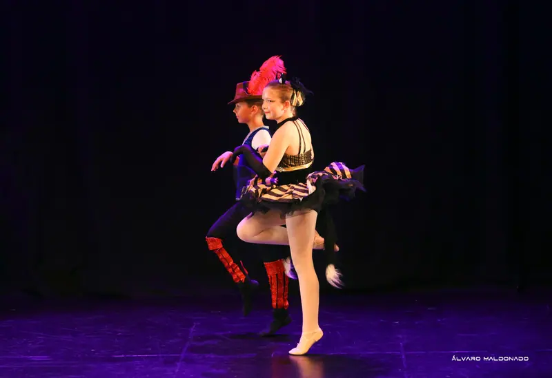 Deux danseurs avancent à l’unisson, l’un en chapeau à plume et bottes rouges, l’autre en tutu rayé avec une queue de chat