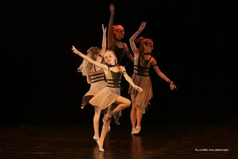 Quatre danseuses en robes kaki à bandes noires évoluent dans une lumière chaude, celle de devant en équilibre sur une jambe