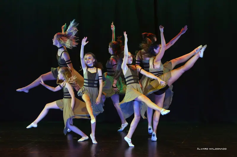 Des danseuses en robes kaki à bandes noires sautent et lancent la jambe, cheveux au vent, dans une pièce contemporaine énergique
