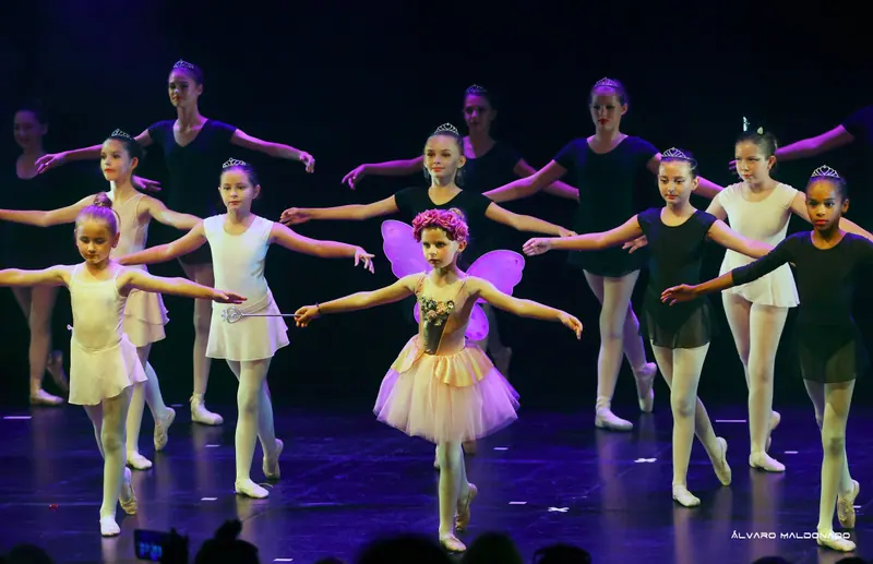 Une petite fée aux ailes violettes et couronne de roses mène des élèves de ballet en justaucorps blancs et noirs avec diadèmes