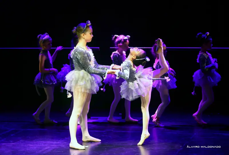 Une danseuse en costume de souris tient les mains d’une autre cambrée en arrière, une jambe levée très haut, la barre derrière