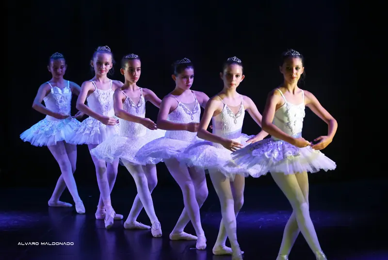 Six filles en tutus classiques blancs et diadèmes forment une diagonale, pieds croisés, sous une lumière lilas