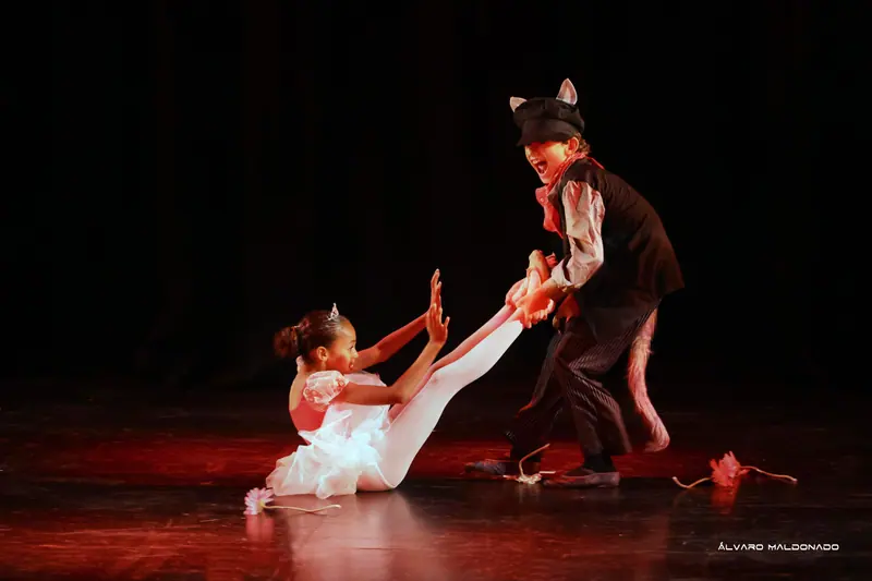 Une fille en tutu blanc et diadème est assise au sol tandis qu’un danseur déguisé en loup avec casquette tire ses jambes
