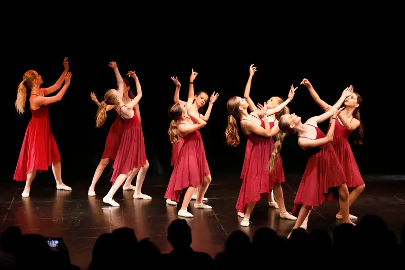 Des danseuses adolescentes en robes bordeaux fluides tendent les bras vers le haut dans une pièce lyrique devant le public