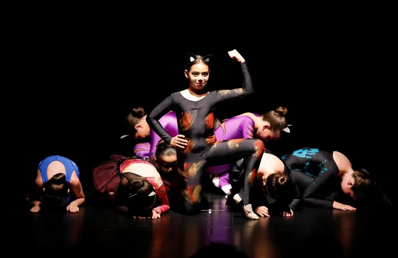 Une fille en combinaison noire peinte avec oreilles de chat montre ses muscles tandis que les autres danseuses s’inclinent au sol