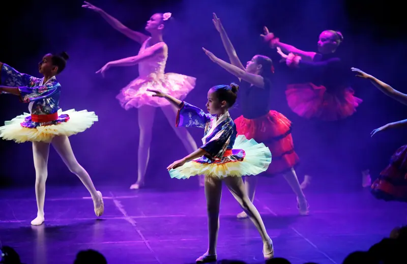 Des filles en hauts kimono bleus fleuris et tutus jaunes dansent sous une lumière violette, des danseuses en tutus rose et rouge derrière