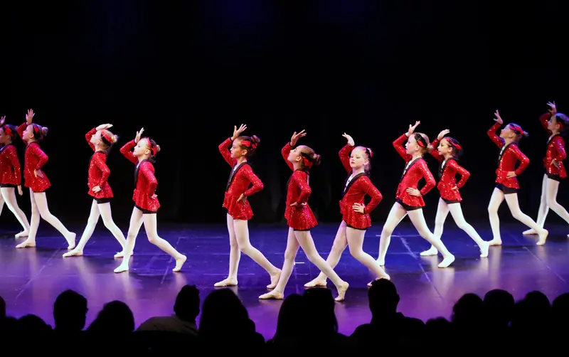 Une file de filles en vestes rouges à paillettes et collants blancs traverse la scène, une main levée au front
