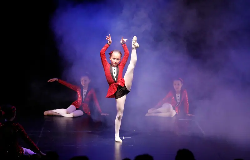Une fille en queue-de-pie rouge à paillettes tient une extension de jambe haute dans une brume violette, deux danseuses assises derrière