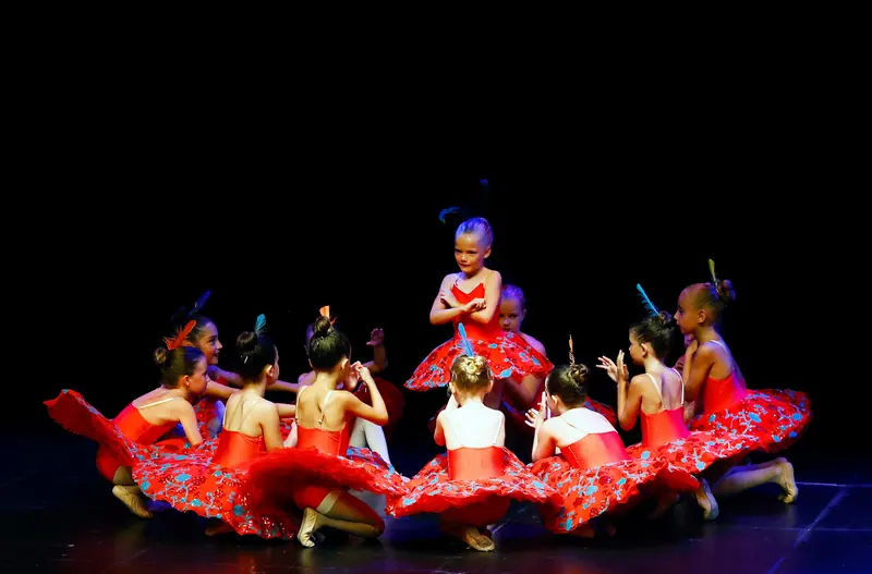 Des filles en tutus rouges fleuris s’agenouillent en cercle autour d’une jeune danseuse debout, bras croisés, une plume dans les cheveux