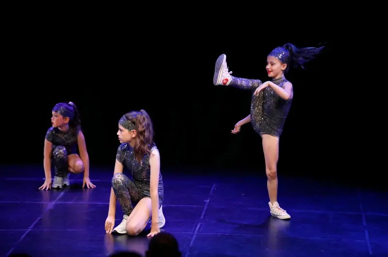 Trois filles en combinaisons grises à paillettes et bandeaux dansent, l’une lance un coup de pied haut en baskets blanches, deux sont accroupies