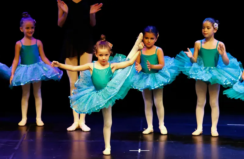 Une fillette en tutu turquoise tient sa jambe levée sur le côté sous le regard de ses camarades en tutus assortis