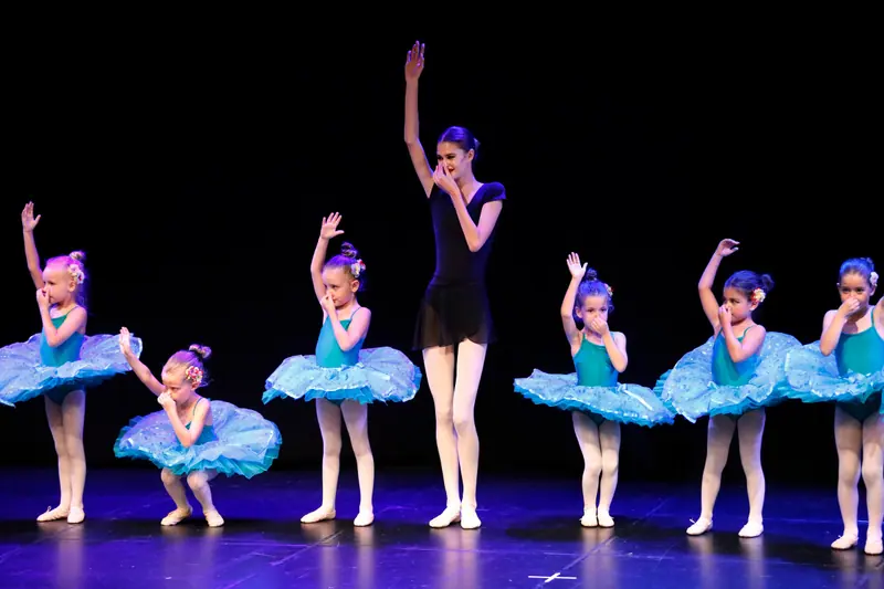De petites ballerines en tutus turquoise se pincent le nez, un bras levé, à côté d’une professeure en justaucorps noir