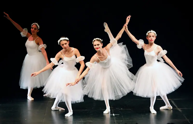 Quatre danseuses en tutus romantiques blancs et couronnes de fleurs se tiennent la main, l’une lève haut la jambe derrière elle