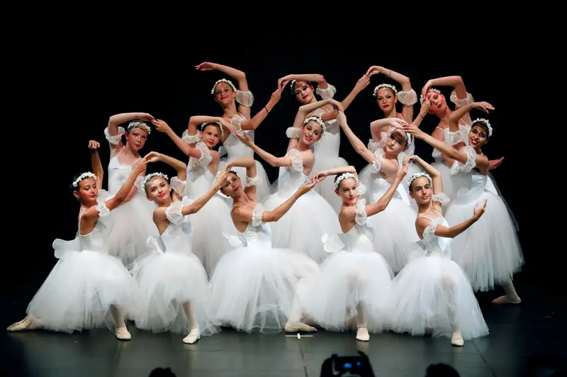 Un grand groupe de danseuses en tutus romantiques blancs et couronnes de fleurs pose les bras arrondis au-dessus de la tête