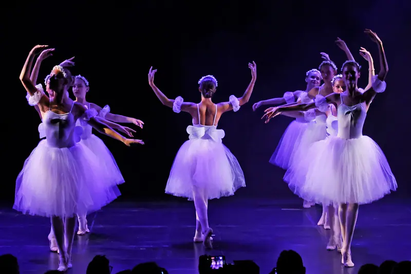 Des danseuses en tutus romantiques blancs à petites ailes lèvent les bras sous une lumière violette, l’une vue de dos
