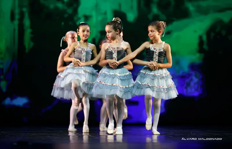 Trois fillettes en velours gris et tutus bleu pâle à volants avancent en dansant main dans la main devant un fond vert