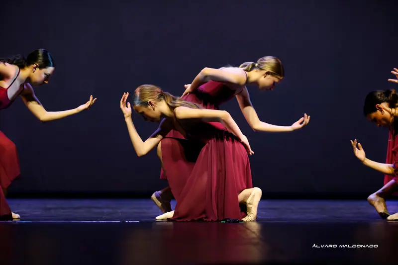 Danseuses en longues robes bordeaux inclinées, mains tendues vers l'avant, l'une à genoux, sur une scène sombre