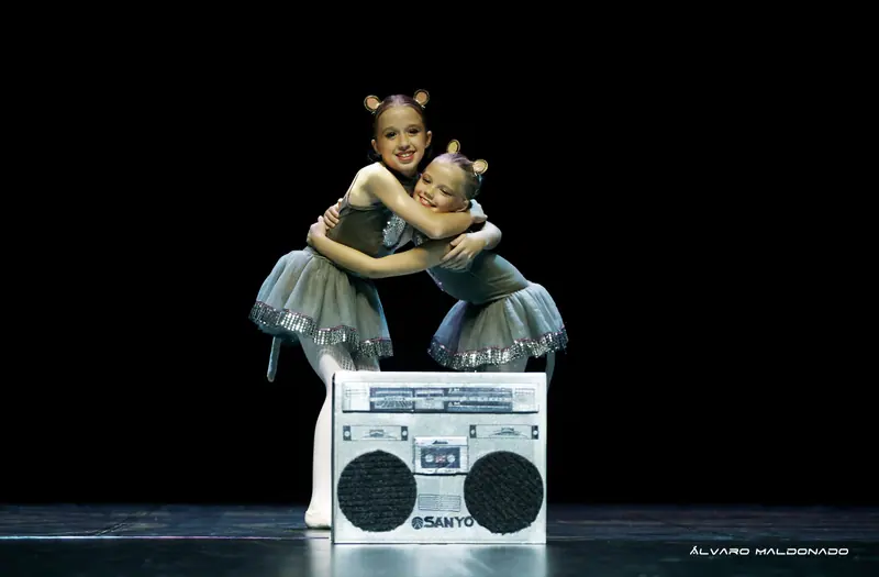 Deux fillettes souriantes en costumes de souris grise à oreilles rondes s'enlacent derrière un grand radiocassette argenté factice
