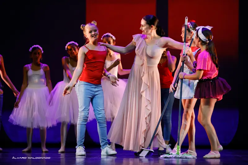 Une professeure en robe rose pâle rit avec des élèves tenant balais et serpillières, des ballerines en tutu blanc derrière
