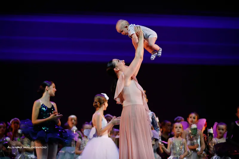 Une femme en robe rose soulève joyeusement un bébé au-dessus de la scène tandis que les élèves costumées applaudissent