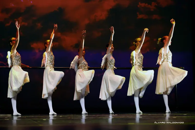 Six fillettes en robes crème et ailes d'ange à plumes lèvent un bras et un genou à la barre devant un ciel rouge nuageux