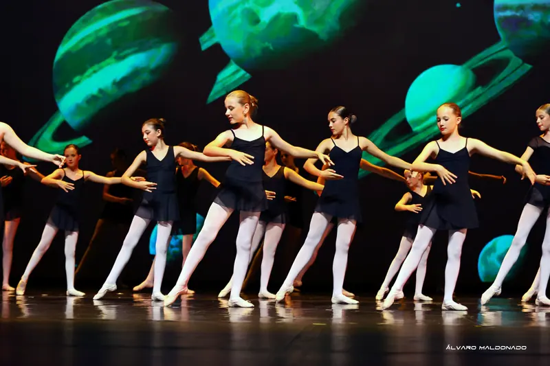 Fillettes en justaucorps et jupettes noirs et collants blancs pointant un pied en avant, bras arrondis, devant une projection de planètes