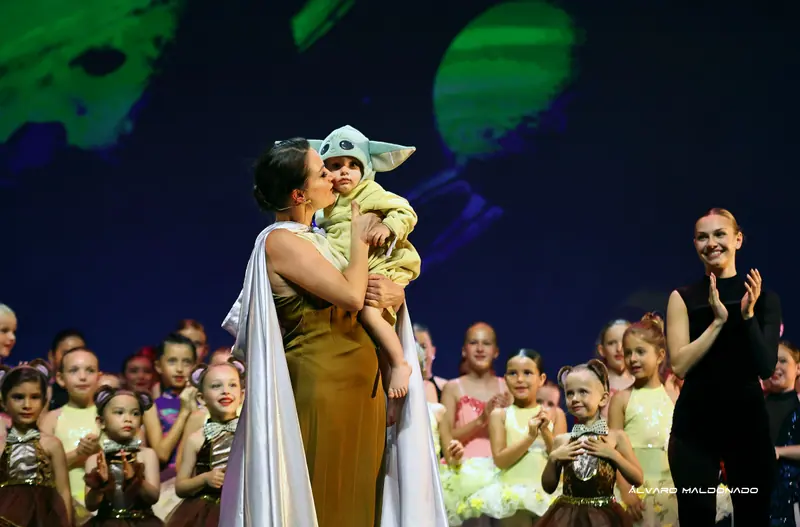 Une femme en robe dorée et cape blanche embrasse un tout-petit en costume vert à grandes oreilles tandis que des fillettes costumées applaudissent