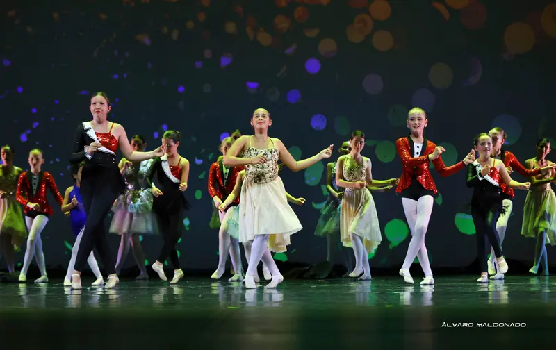 Un grand groupe aux costumes variés, des vestes rouges à sequins à une robe dorée, danse le final devant des lumières colorées floues