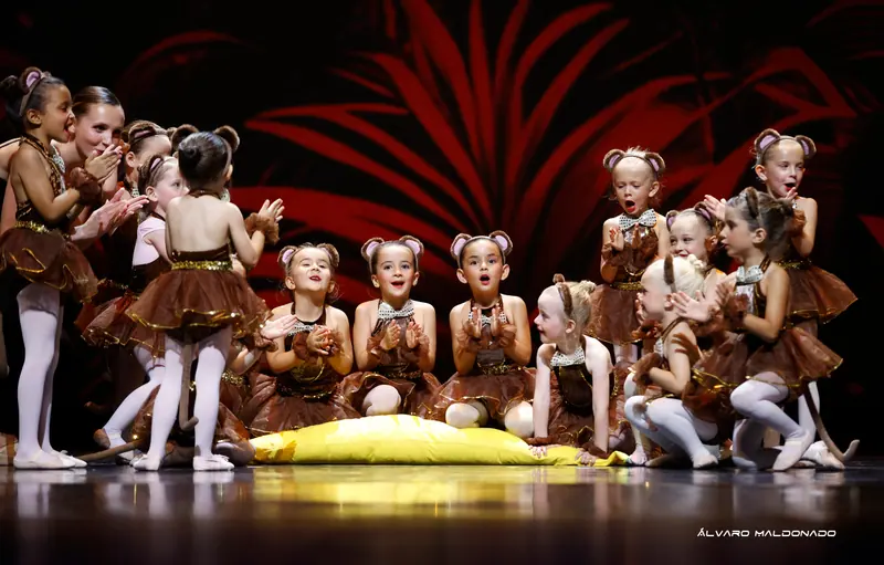 Petites filles en costumes de singe marron à oreilles rondes, agenouillées autour d'un grand coussin jaune, applaudissent émerveillées