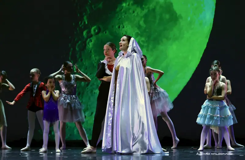 Une interprète en cape de satin blanc à capuche chante avec un micro serre-tête parmi de jeunes danseuses devant une immense lune verte