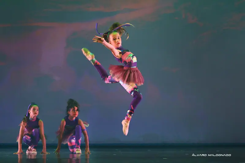 Une fillette en combinaison à motifs colorés et tutu bordeaux saute haut tandis que deux danseuses sont accroupies en dessous