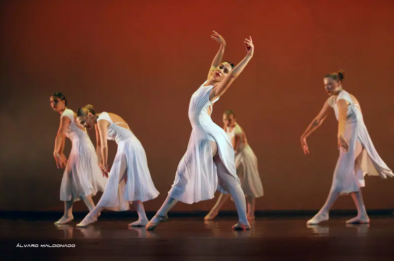 Danseuses en robes blanches fluides traversant la scène, celle du centre cambrée bras levés, sous une chaude lumière orangée