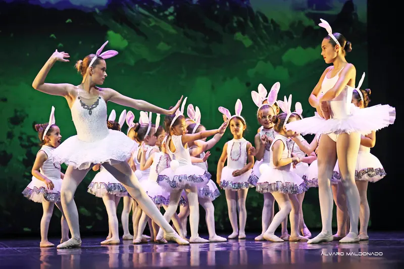 Fillettes en tutus blancs et oreilles de lapin imitent deux danseuses plus âgées en blanc devant un décor de montagnes vertes