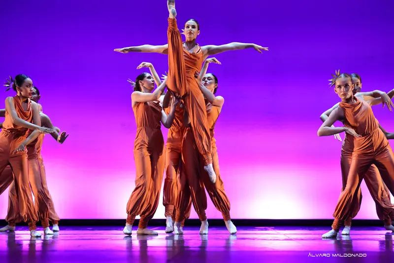 Une danseuse en combinaison cuivrée est soulevée par son groupe, une jambe tendue vers le haut, sous une lumière violette et rose