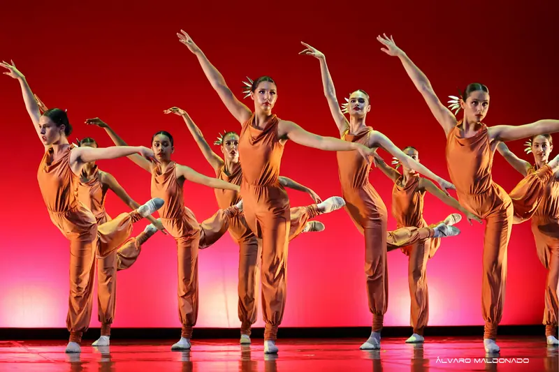 Danseuses adolescentes en combinaisons cuivrées levant une jambe en arrière, bras tendus, devant un fond rouge vif