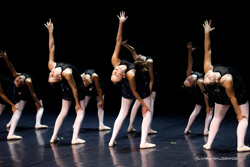 Danseuses en robes sombres à paillettes et collants roses se penchent de côté, un bras tendu vers le haut, sur scène sombre