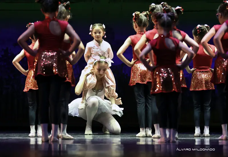 Une fillette en rose est assise sur les épaules d'une danseuse agenouillée en tutu blanc, entourée de filles en paillettes rouges