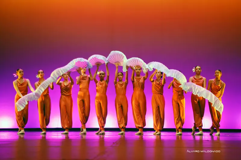 Une ligne de danseuses en combinaisons orange lève des éventails de soie blanche, formant une vague devant un fond aux tons de coucher de soleil