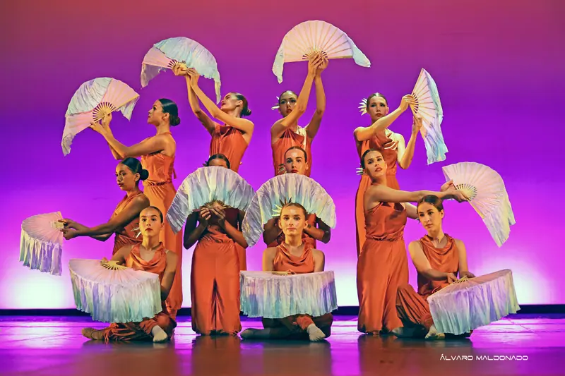 Danseuses en combinaisons orange posent en étages avec de grands éventails de soie blanche, certaines à genoux, devant un fond magenta